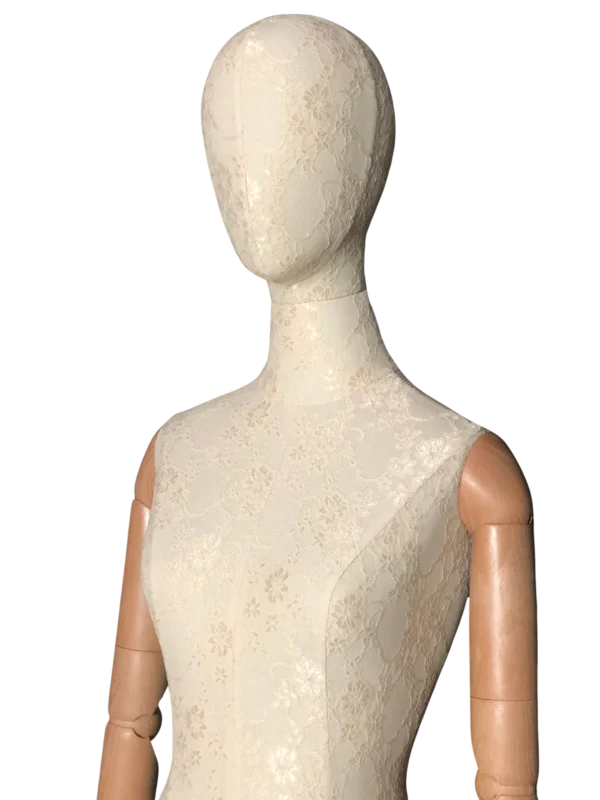 Pandangan dekat depan badan peragawati wanita XINTU, dilitupi fabrik renda putih, dengan kepala abstrak dan lengan kayu yang diartikulasikan.